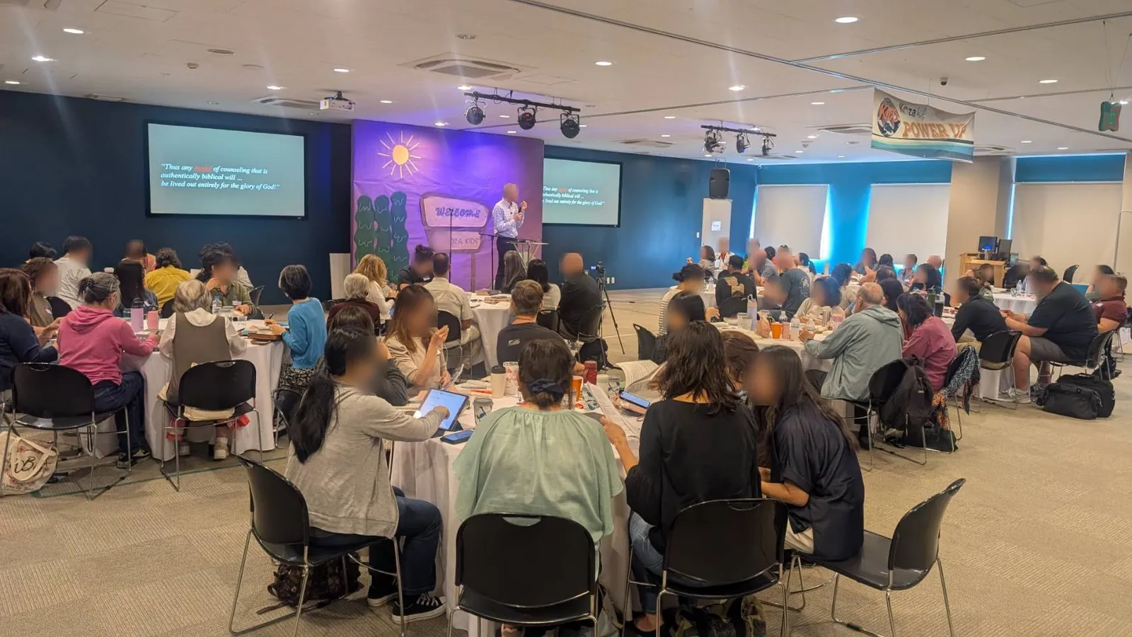 Attendees at round tables during a JBCA training seminar, with a speaker presenting between two projection screens
